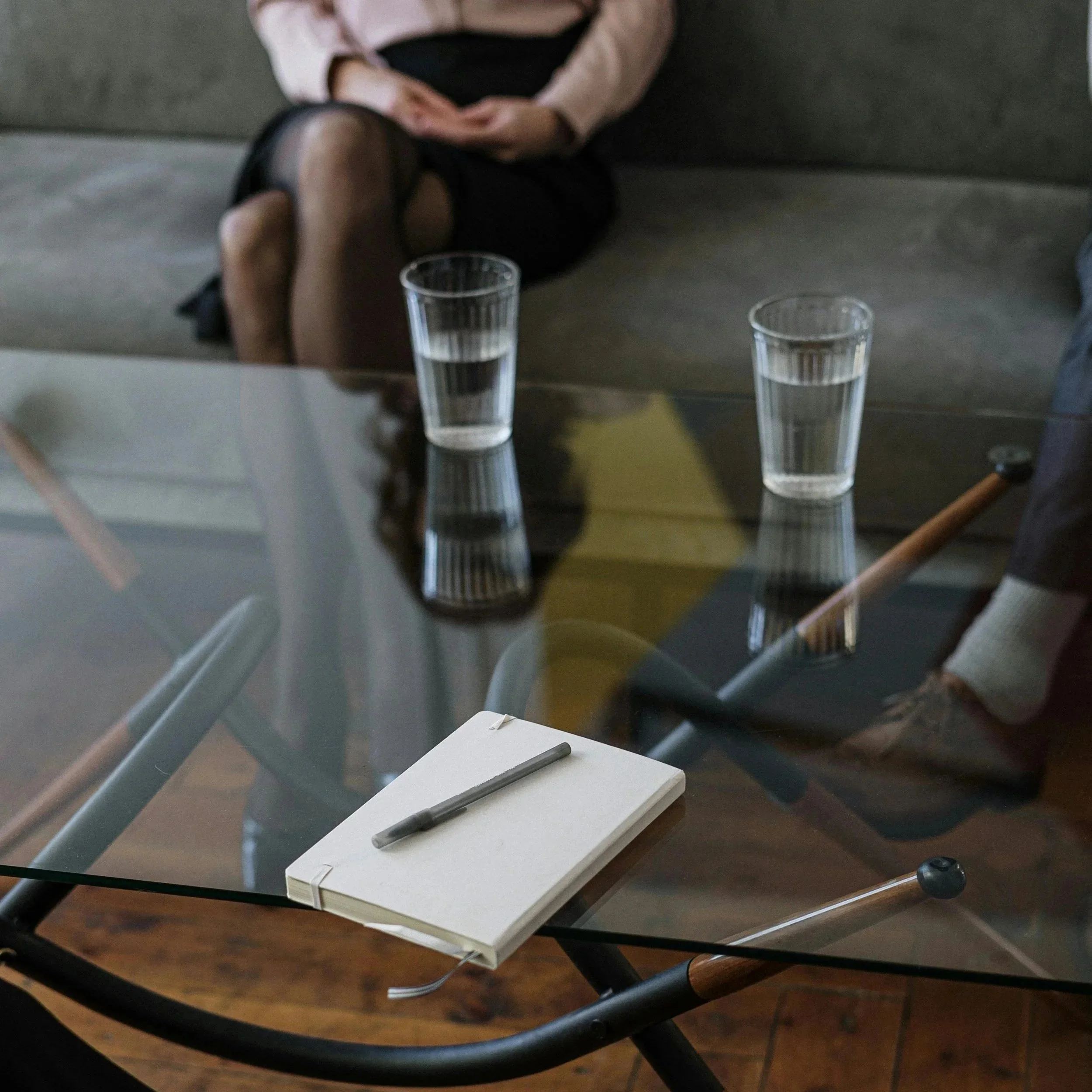 A glass-top table with a notebook and pen, two glasses of water, and reflections on the surface. In the background, a woman with crossed legs in black stockings and a pink sweater sits on a couch.