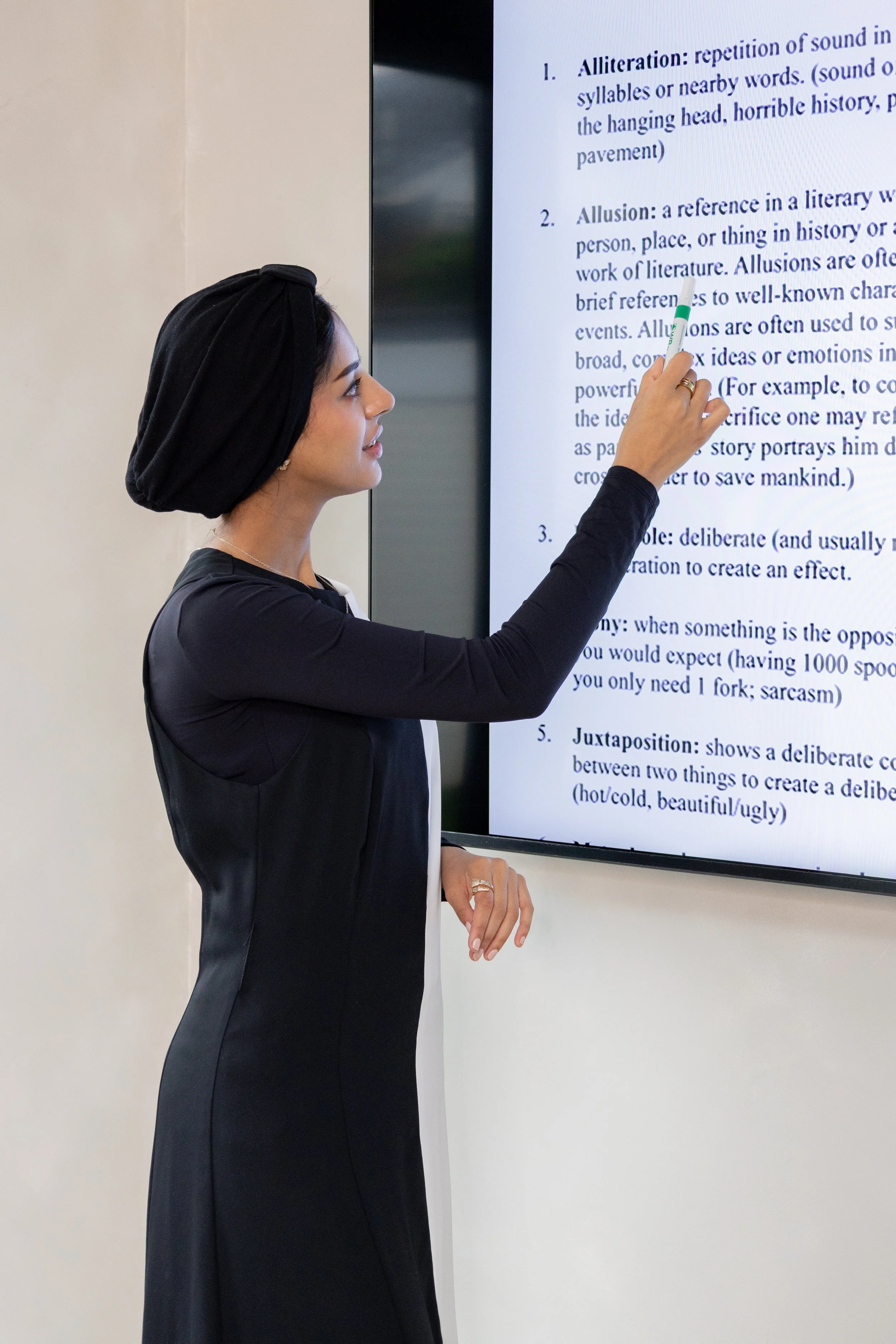 A woman wearing a black headscarf and a black dress points at a large screen displaying a text document with a green marker.