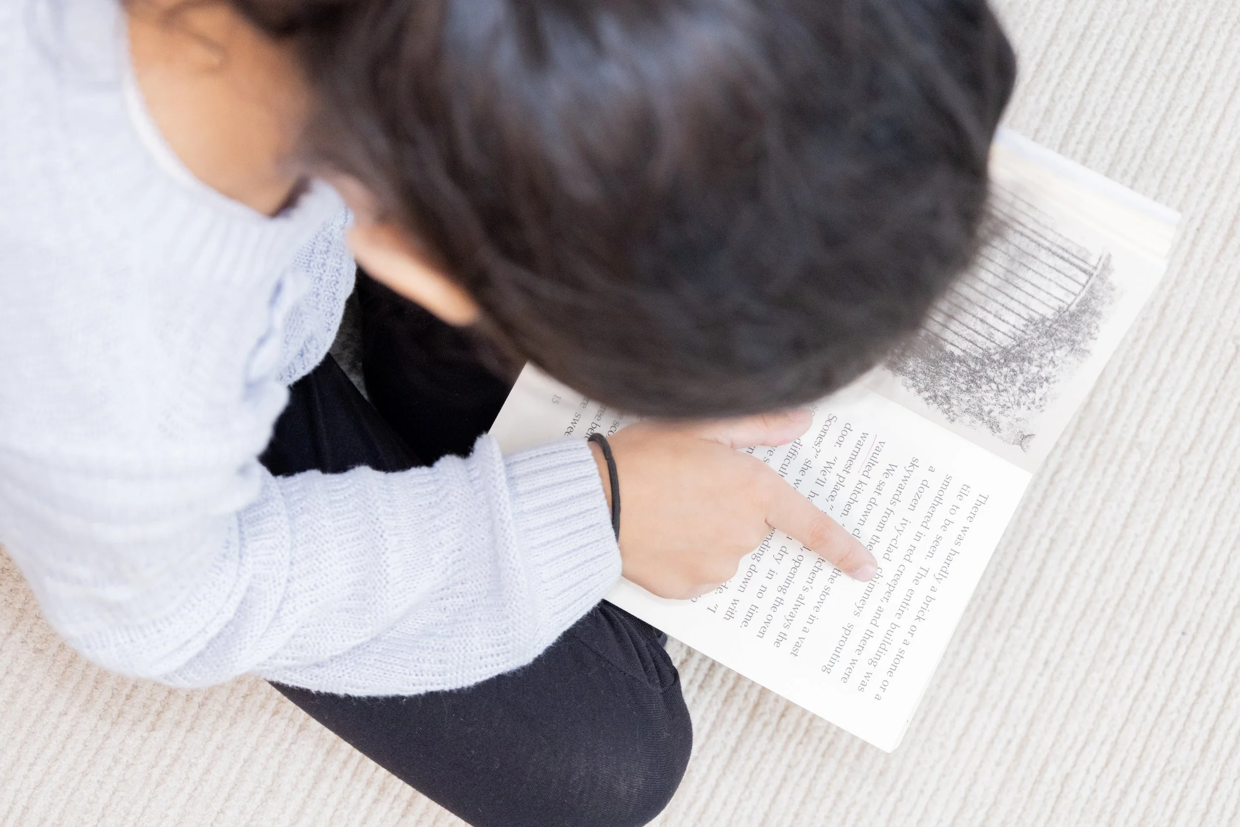 Person sitting on the floor reading a book with a grayscale image of a landscape or cityscape on one page.