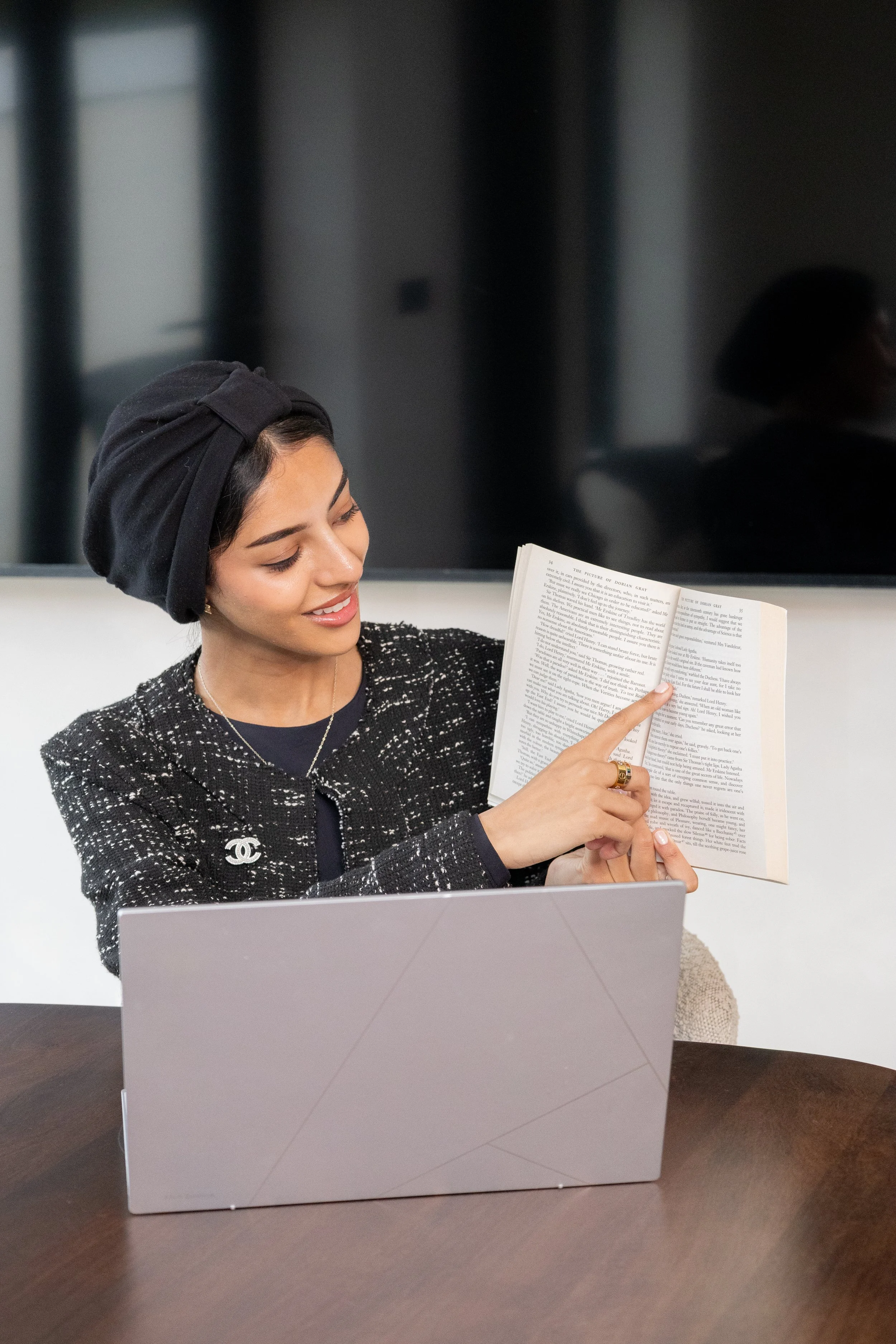 A woman with a black headscarf and a black and white tweed jacket sitting at a table, reading a book and pointing at the text. There is a laptop in front of her.