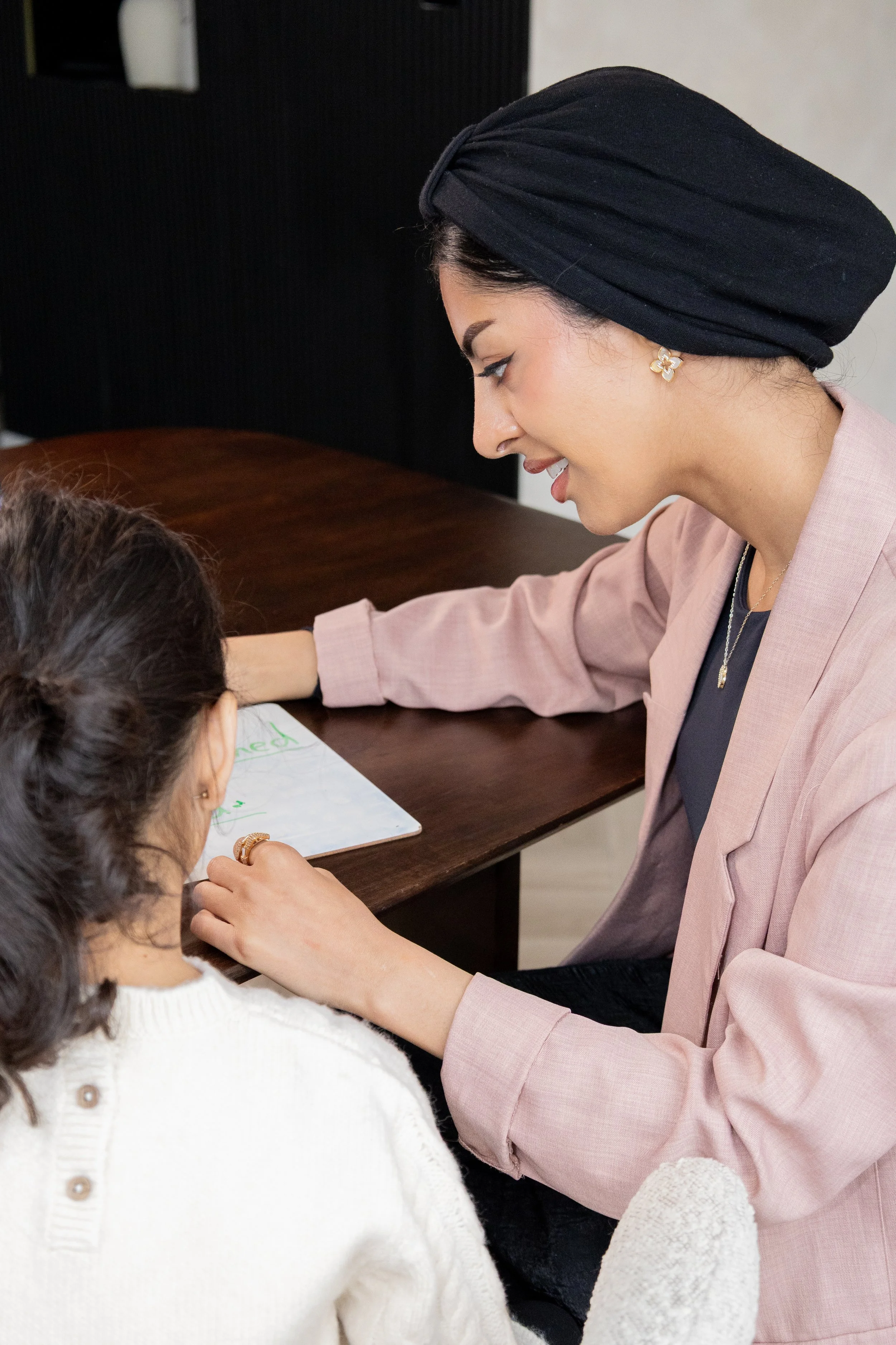 A woman with a black headwrap, earrings, and a pink blazer smiling and helping a young girl with her homework at a desk.