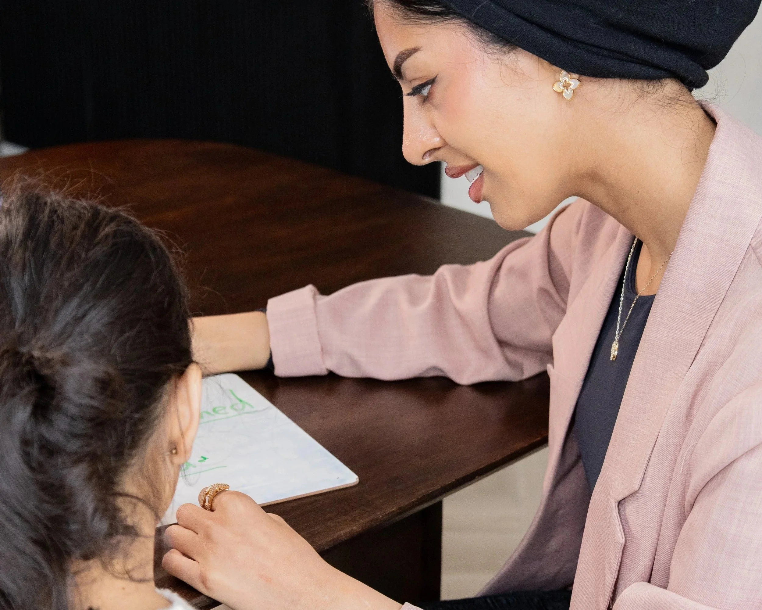 A woman with dark hair, wearing a black head covering and a pink blazer, smiling and leaning over a table, looking at a young girl with dark hair tied back, who is sitting on the other side of the table with a white paper in front of her.