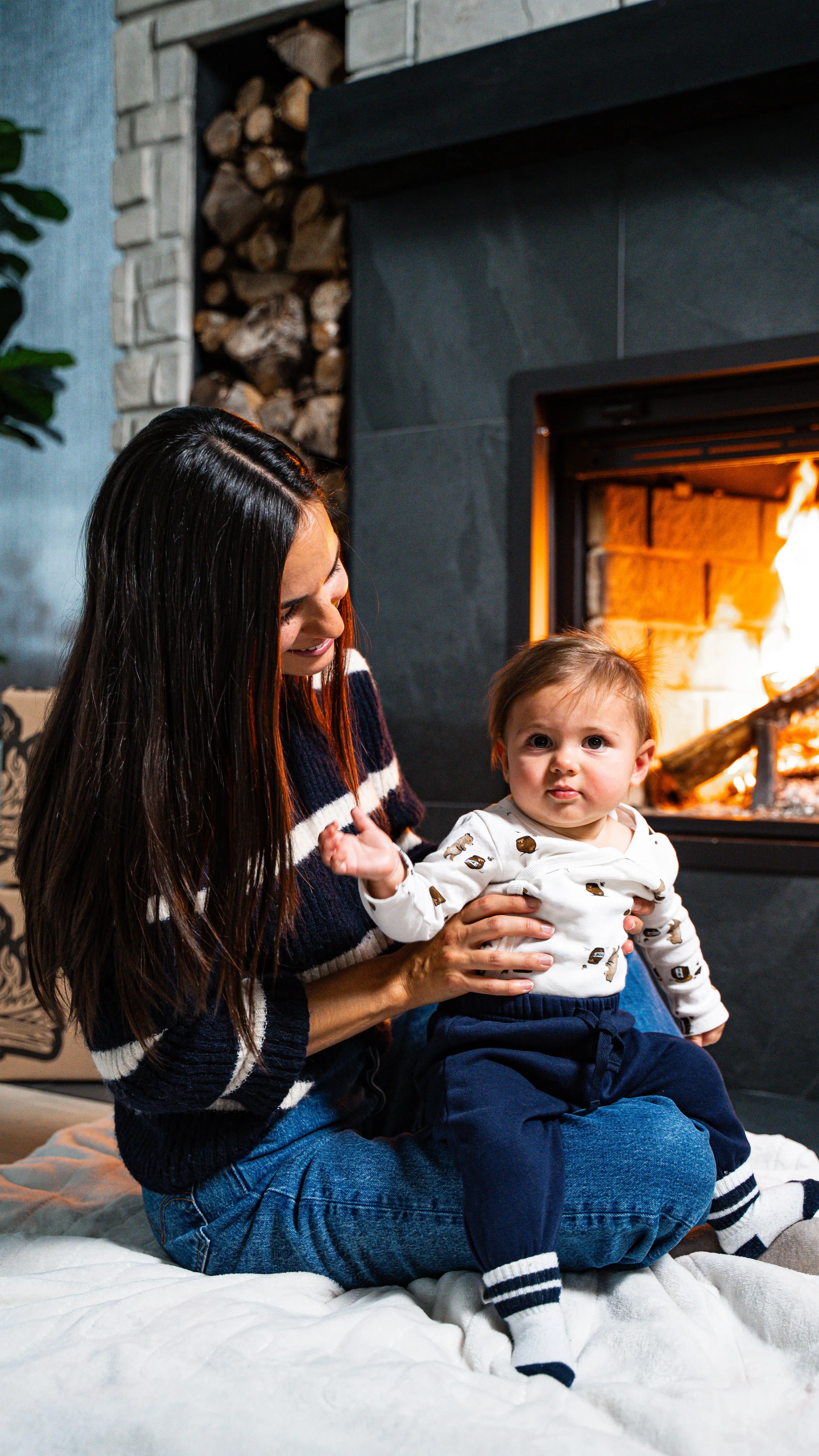 Une femme et un jeune garçon assis près d'une cheminée en train de faire un feu, la femme sourit et regarde le garçon.
