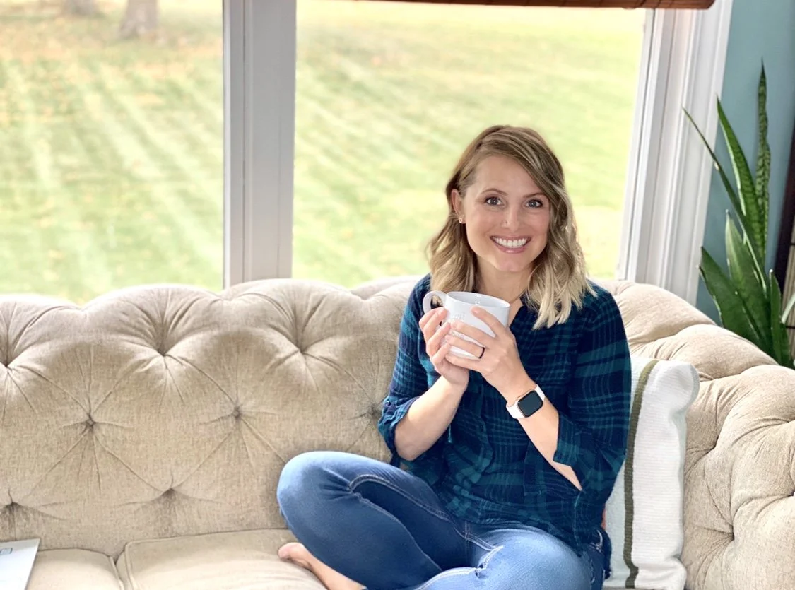 A woman with blonde hair smiling and holding a white mug, sitting cross-legged on a tufted beige sofa in front of a large window with green outdoor scenery.