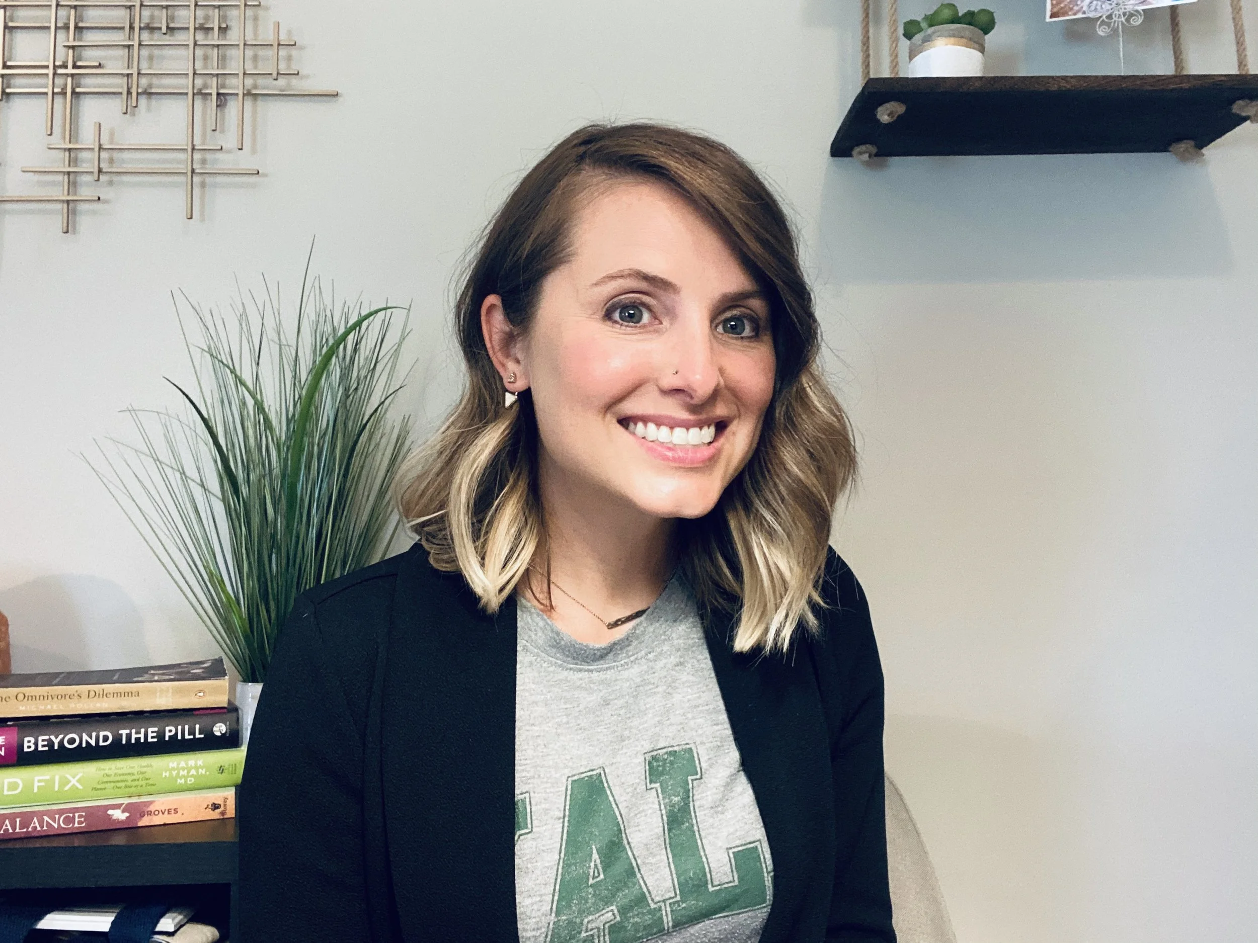 A woman with shoulder-length wavy brown hair, wearing a gray T-shirt with green letters and a black blazer, smiling while sitting in a modern living room with plants and books in the background.