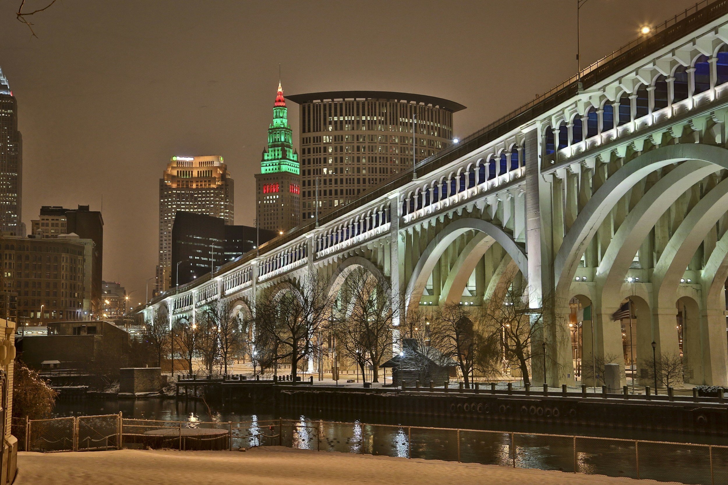 Nighttime cityscape of Cleveland, Ohio, featuring the illuminated Cleveland Flatiron Building with colorful tower lights, the historic Major League Baseball Park, and a lit arched bridge over a snow-covered river, with a cloudy sky in the background.