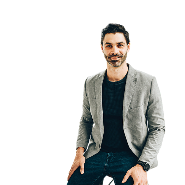 A man with dark hair and a beard wearing a gray blazer and black shirt, smiling and sitting with one hand resting on his thigh against a colorful background.