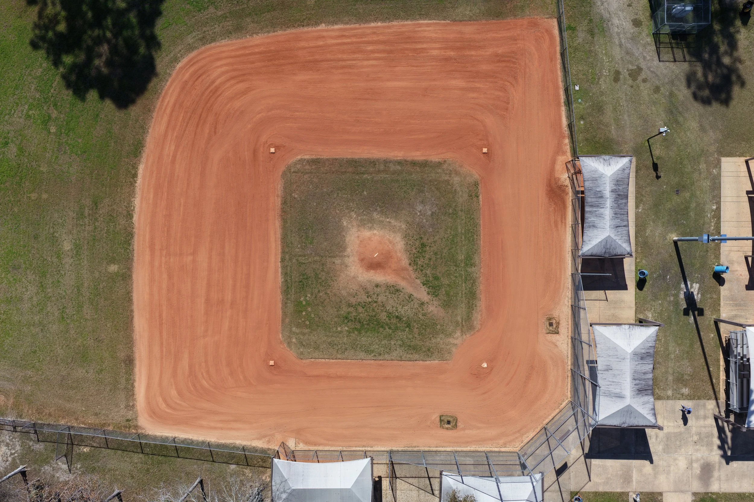 Aerial view of a baseball or softball field with a dirt infield and a grass outfield, surrounded by a chain-link fence, with structures and equipment nearby.