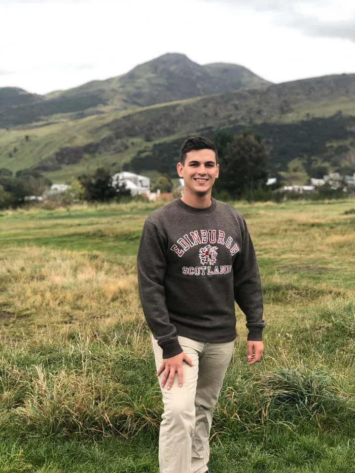 A young man standing in a grassy field with mountains in the background, wearing a gray sweater that says 'Edinburgh Scotland' and beige pants.