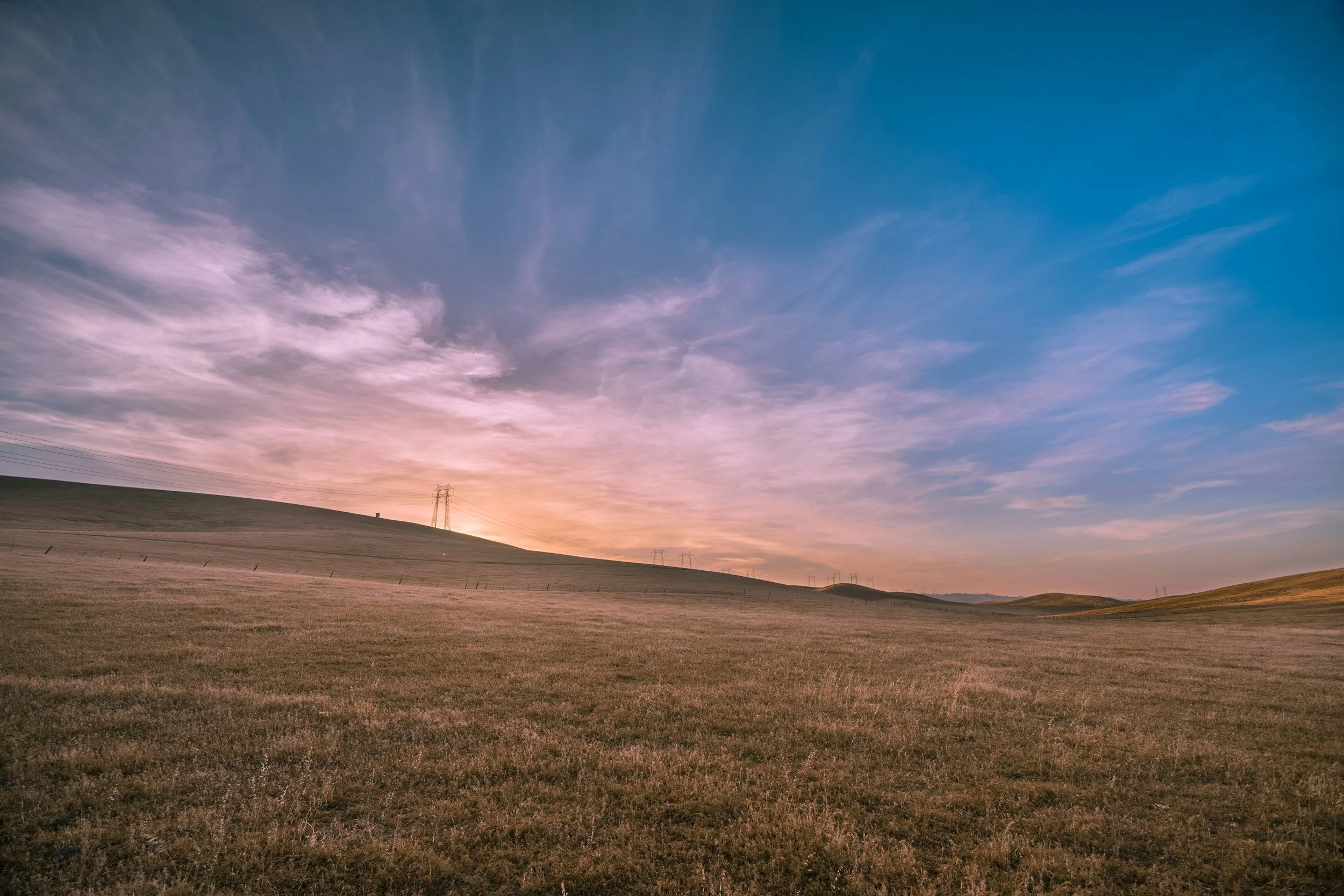 Open field with grass, rolling hills, power lines and towers at sunset, partly cloudy sky with pink and blue hues