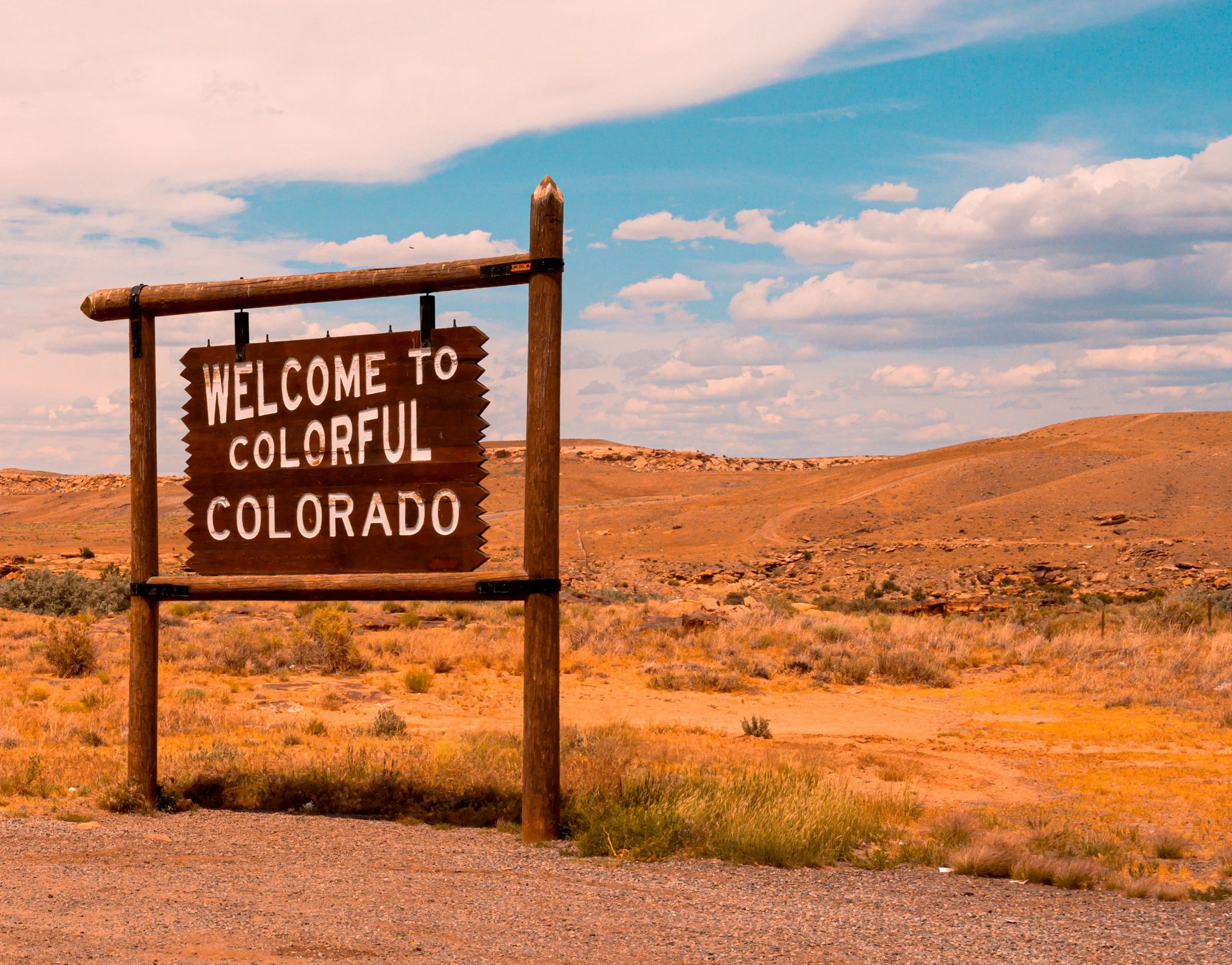 A wooden sign reading 'Welcome to Colorful Colorado' in a desert landscape with rolling hills and a partly cloudy sky.