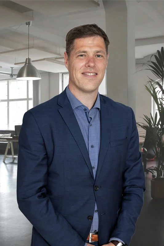 Allan i blå habitus og blå skjorte står i et moderne kontor med store vinduer og grønne planter.