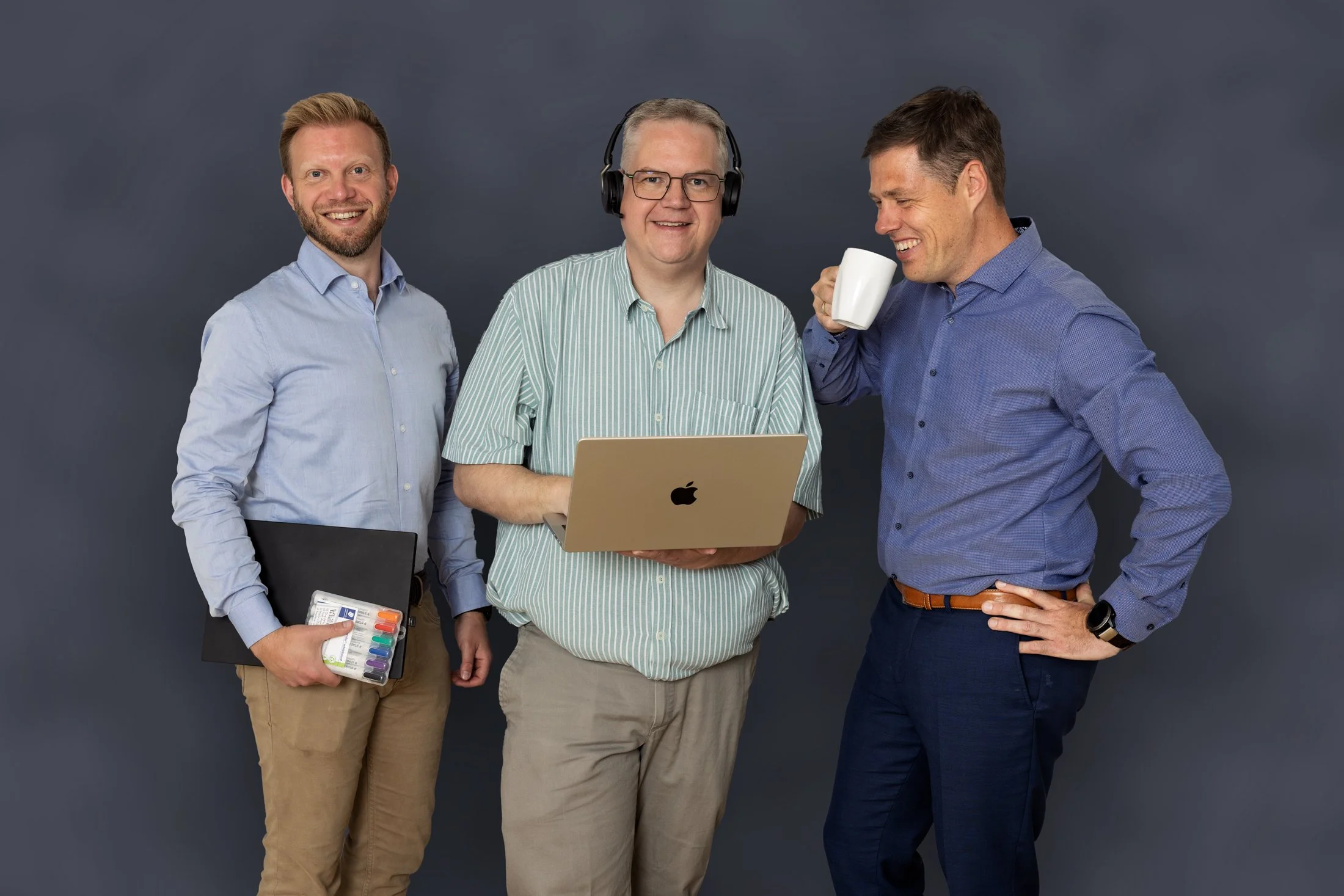 De tre ejer i kontorhurtigt tøj, der står sammen mod en væg, hvoraf den midterste har høretelefoner og bruger en bærbar computer, den højre har en kaffekop, og den venstre holder dokumenter.
