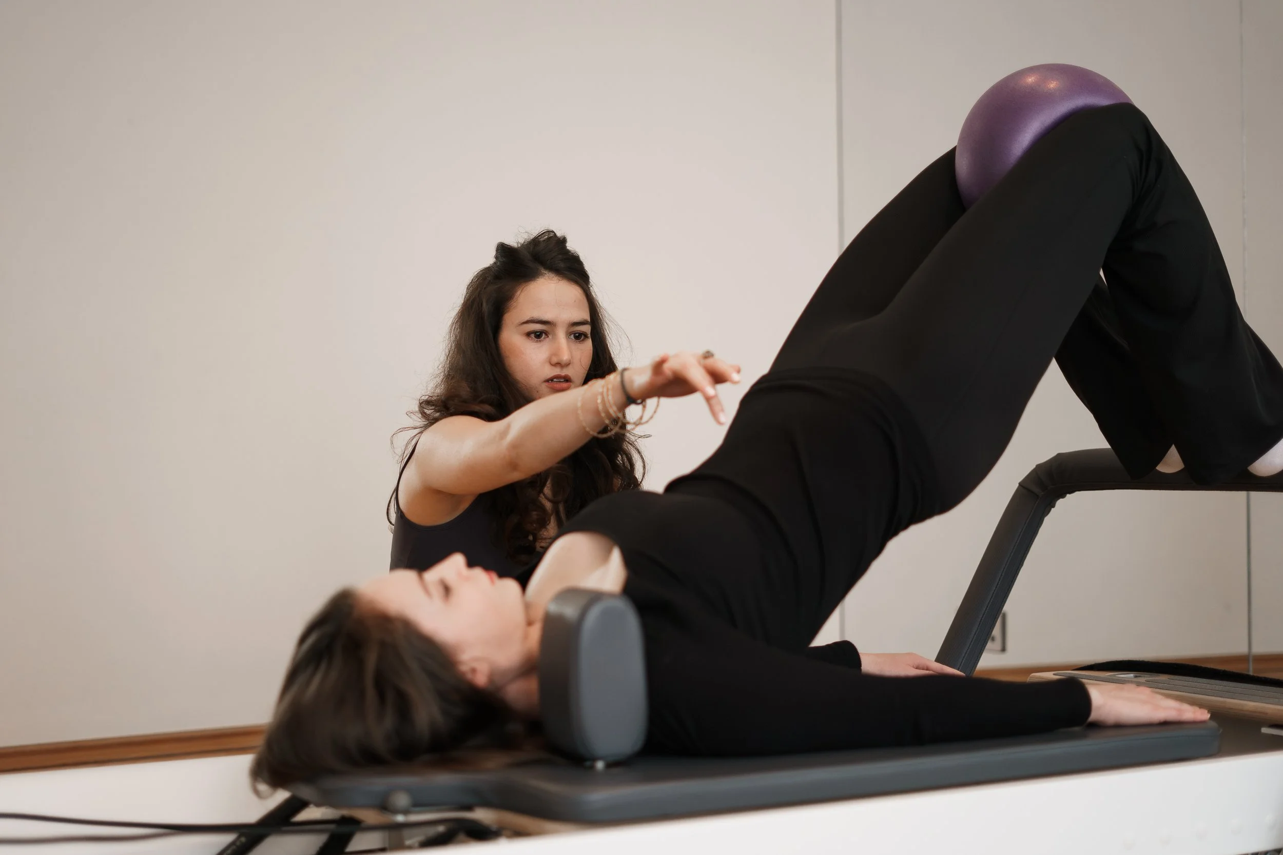 A woman with brown hair lying on a workout bench with her knees bent, wearing black leggings and a black top, while a trainer with dark brown hair observing and guiding her during a Pilates or physical therapy session in a fitness studio.