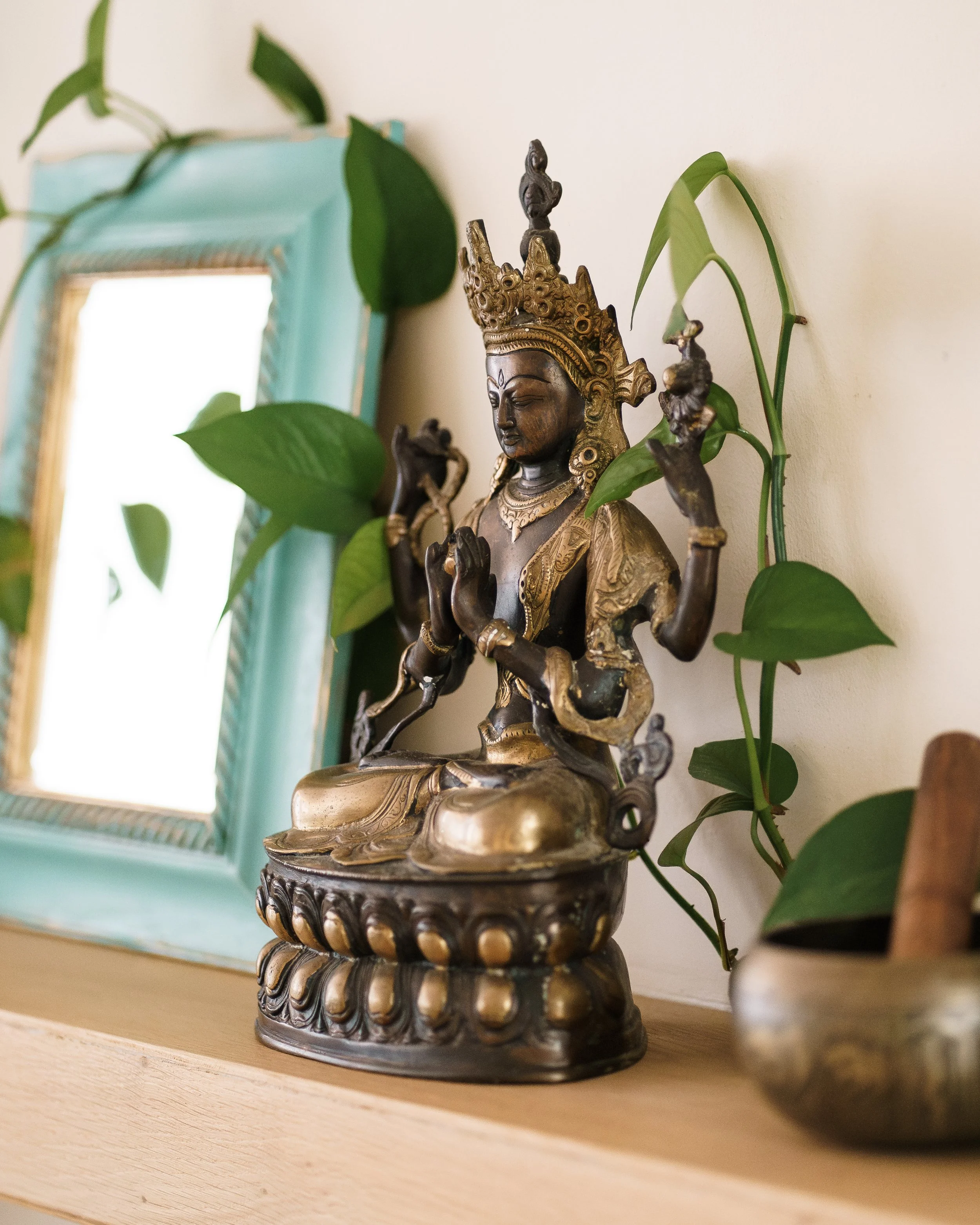 A decorative metal statue of a multi-armed deity, surrounded by green plant leaves, placed on a wooden surface with a framed mirror in the background.