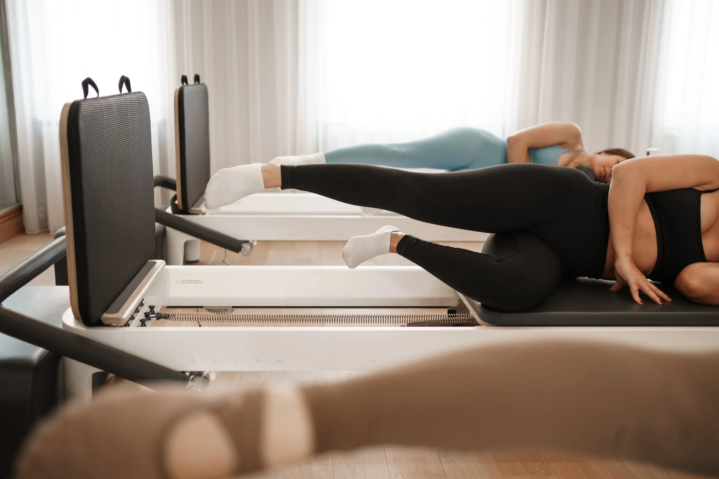 A woman in black leggings and a black sports top performs leg lifts on a Pilates reformer machine in a gym, with another woman in blue lying on a mat in the background.