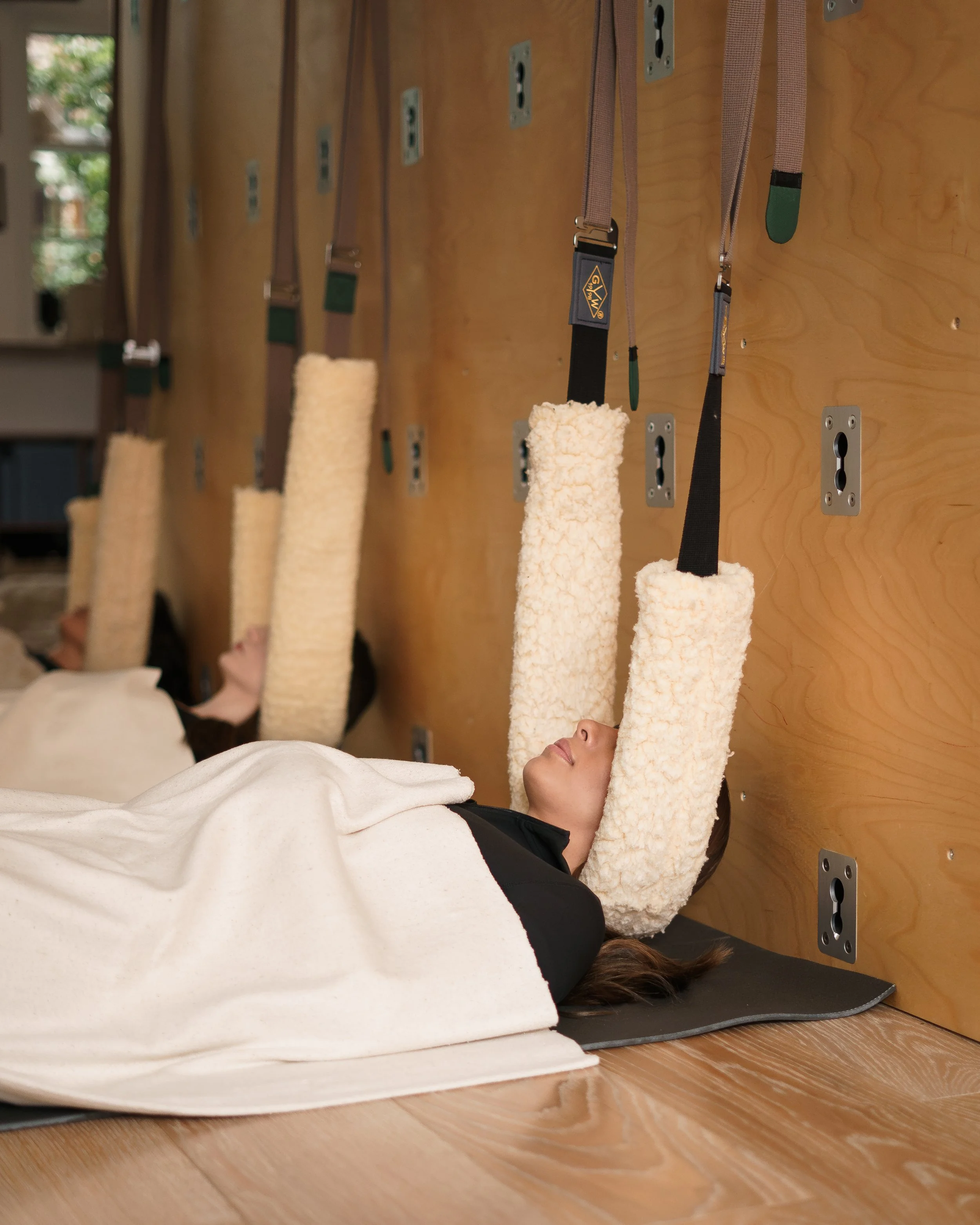 A person lying on a mat while holding onto a suspended yoga or therapy strap attached to a wall unit with multiple straps.