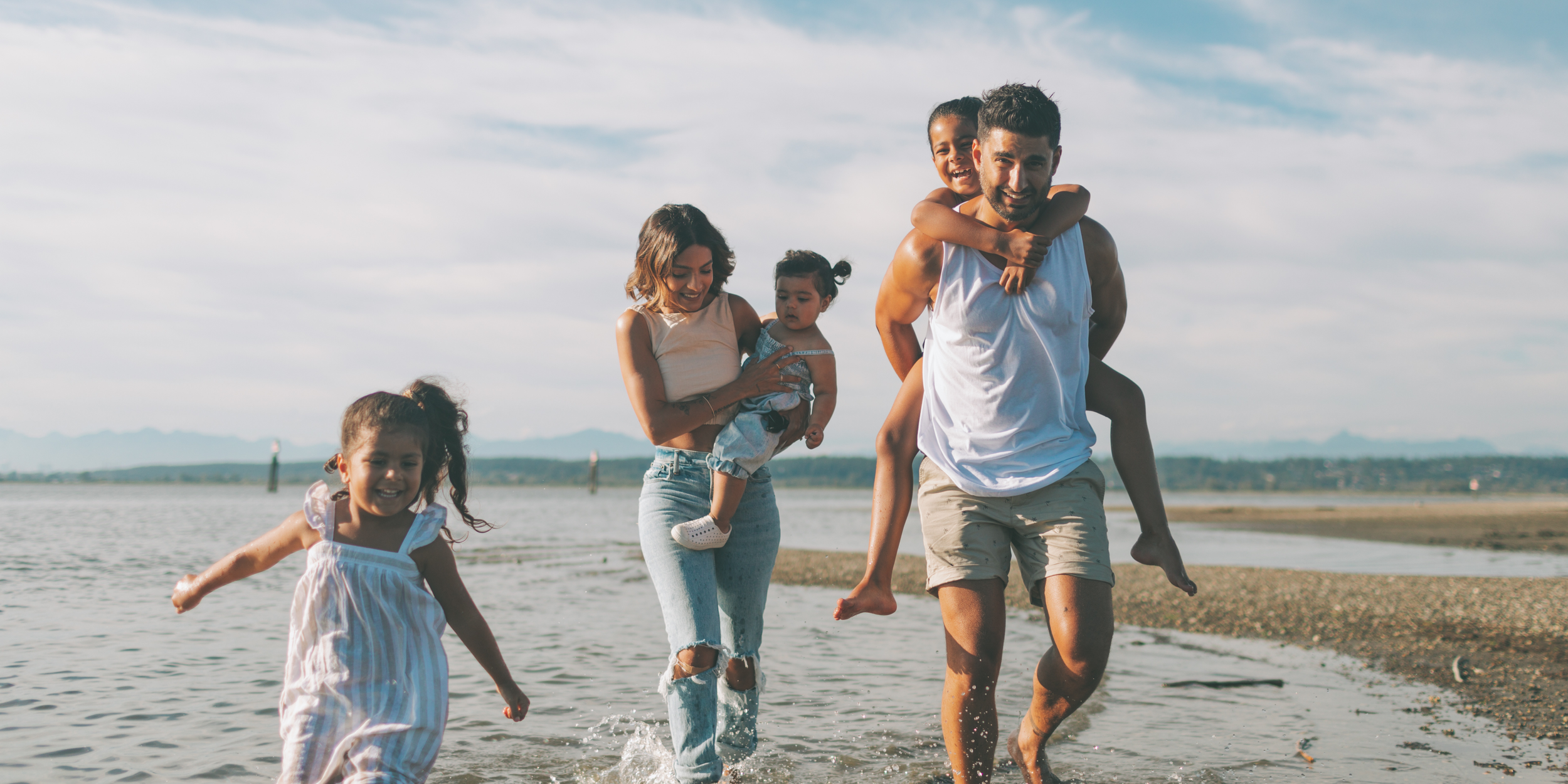 Naples, FL - Family of five, including two young girls and two parents, enjoying a walk along the beach with shallow water, distant mountains, and partly cloudy sky in the background.