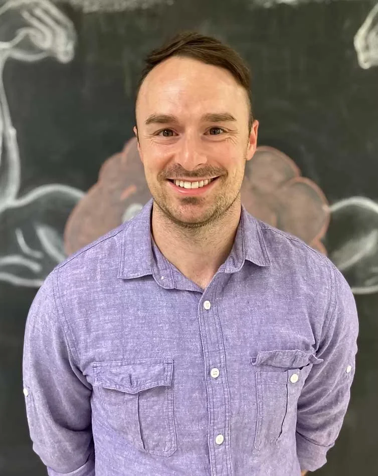 A smiling man with short brown hair wearing a light purple button-up shirt. There is a chalkboard with a faint drawing of a brain in the background.