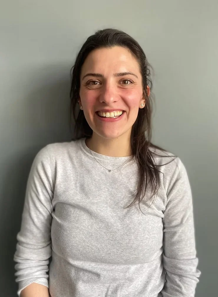 A young woman with dark hair, wearing a light gray long-sleeve shirt, smiling and standing against a plain light gray wall.