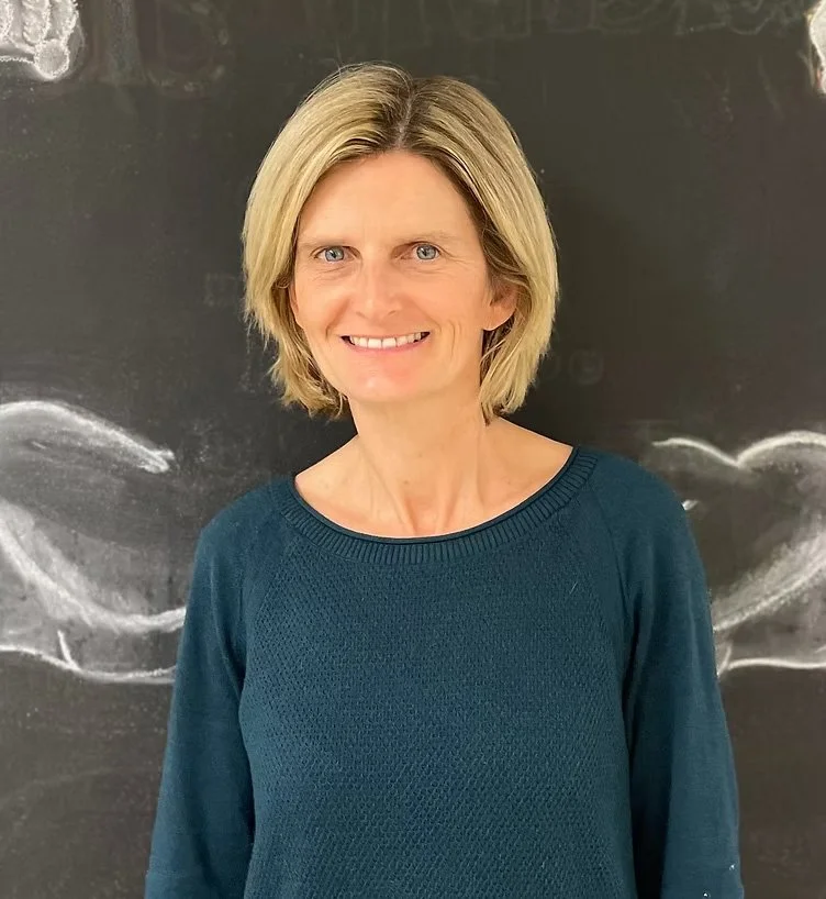 A woman with blonde hair wearing a blue sweater, smiling, standing in front of a blackboard with chalk drawings.