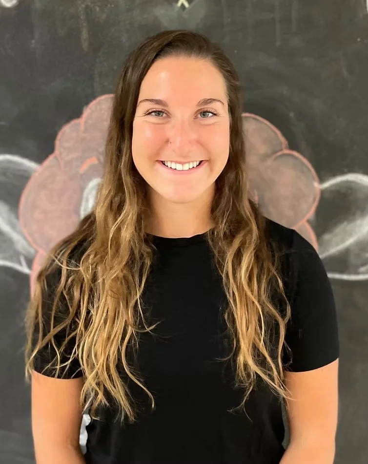 A young woman with long wavy blond hair, smiling, wearing a black T-shirt, standing in front of a blackboard with drawings of a flower and chalk lines.