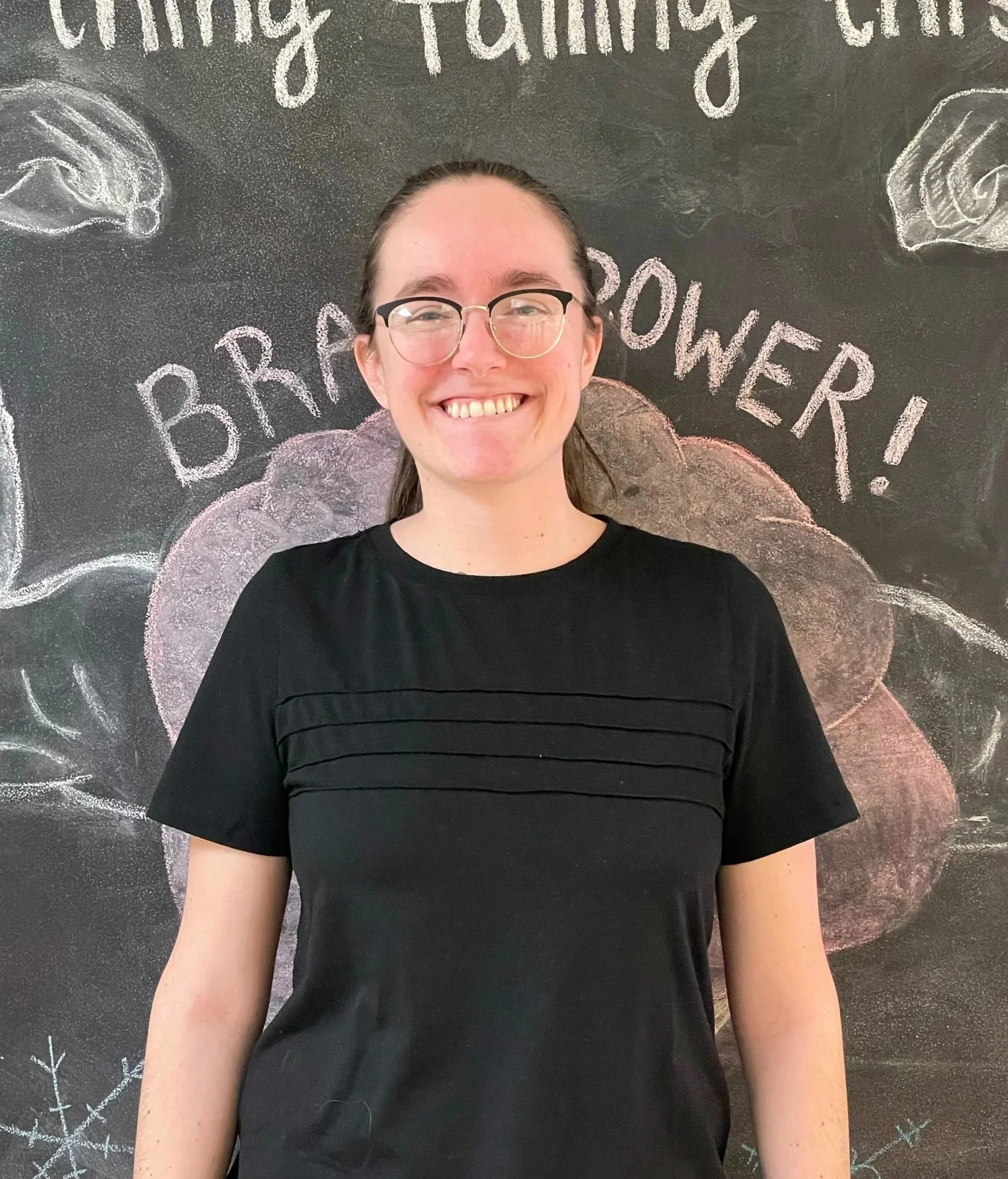 A smiling woman wearing glasses and a black shirt standing in front of a chalkboard wall decorated with festive drawings and the words 'BRRR! POWER!' and 'Warm Fuzzy Feeling' written on it.