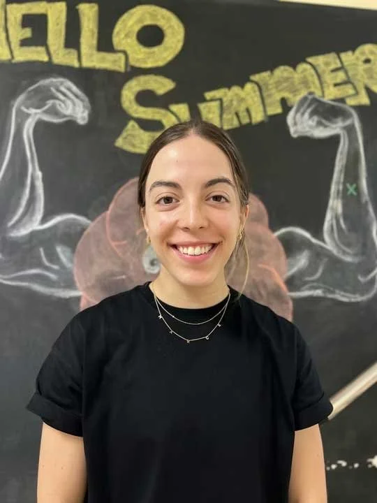 A young woman with brown hair smiling in front of a chalkboard with a chalk drawing of a brain and muscular arms, and the words 'HELLO SUMMER' written in yellow.