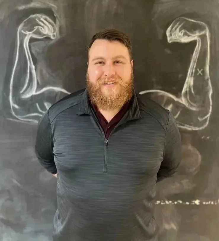 A man with a beard and short hair standing in front of a chalkboard with white chalk drawings of flexed biceps.