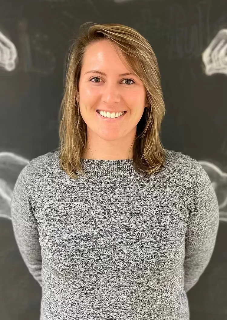A woman with shoulder-length light brown hair smiling, wearing a gray speckled top, standing in front of a dark gray chalkboard with drawings.