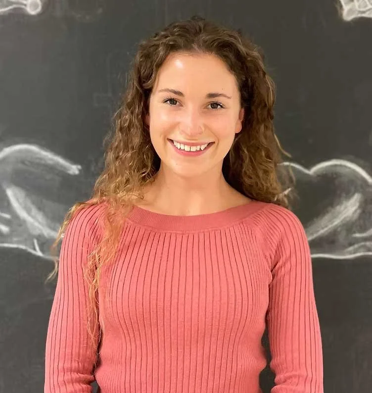 A young woman with curly brown hair smiling, wearing a pink ribbed long-sleeve top, standing in front of a black chalkboard with a drawing of a whale.