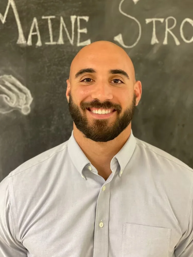A smiling man with a beard and a shaved head, wearing a light-colored button-up shirt, standing in front of a chalkboard with writing and drawings.