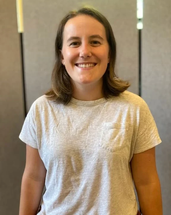 A young woman with shoulder-length brown hair, smiling, wearing a light gray t-shirt, standing indoors in front of a gray partition.