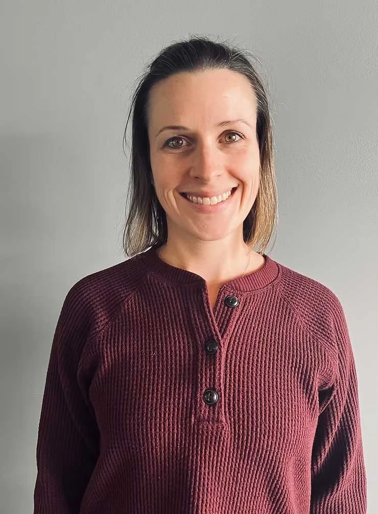 A woman with shoulder-length brown hair and fair skin smiling, wearing a maroon, ribbed, long-sleeve shirt with black buttons, standing against a plain gray wall.
