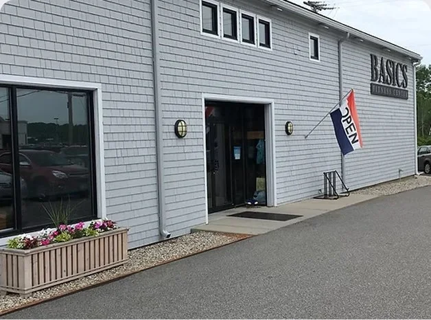 Exterior of a building with a sign that reads 'BASICS FITNESS CENTER'. There is an open door with a mat in front, and a flag that says 'OPEN' near the door. A flower planter is beside the door.