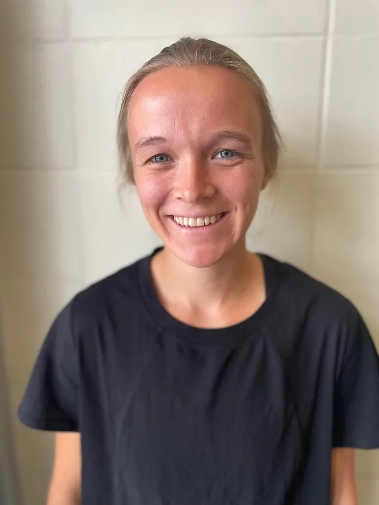 A woman with blonde hair, blue eyes, and a broad smile, wearing a black T-shirt, standing against a light-colored wall.
