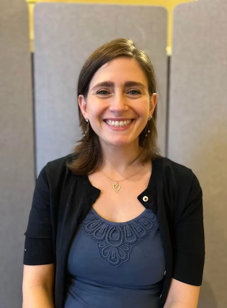 A smiling woman with shoulder-length brown hair, wearing a dark blue top with lace detailing, a black cardigan, and jewelry, standing in front of a gray partition.