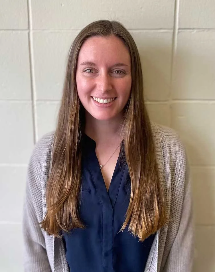 A young woman with long brown hair and a smile, wearing a navy blue shirt and a gray cardigan, standing against a beige cinder block wall.