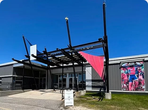 An outdoor art installation with black metal structures and a pink cloth, in front of a building with large windows and a colorful mural, under a clear blue sky.
