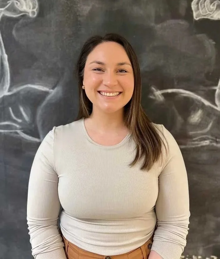 A young woman with long brown hair smiling, wearing a beige long-sleeve shirt and beige pants, standing in front of a chalkboard with chalk doodles.