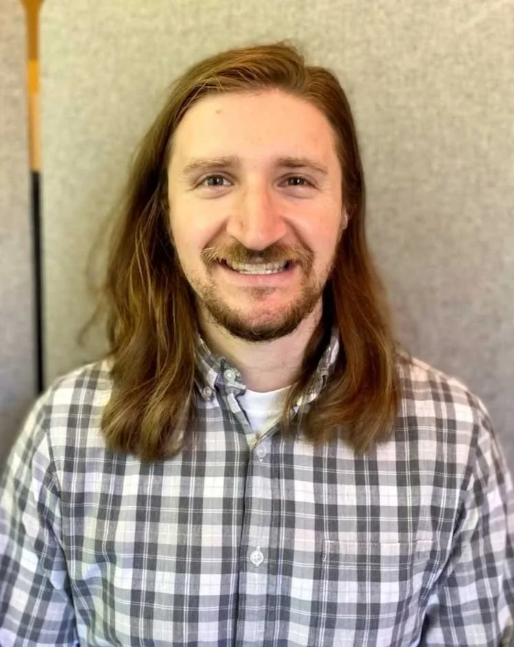 A man with long brown hair, a beard, and a mustache, smiling, wearing a plaid button-up shirt, standing in front of a neutral background.