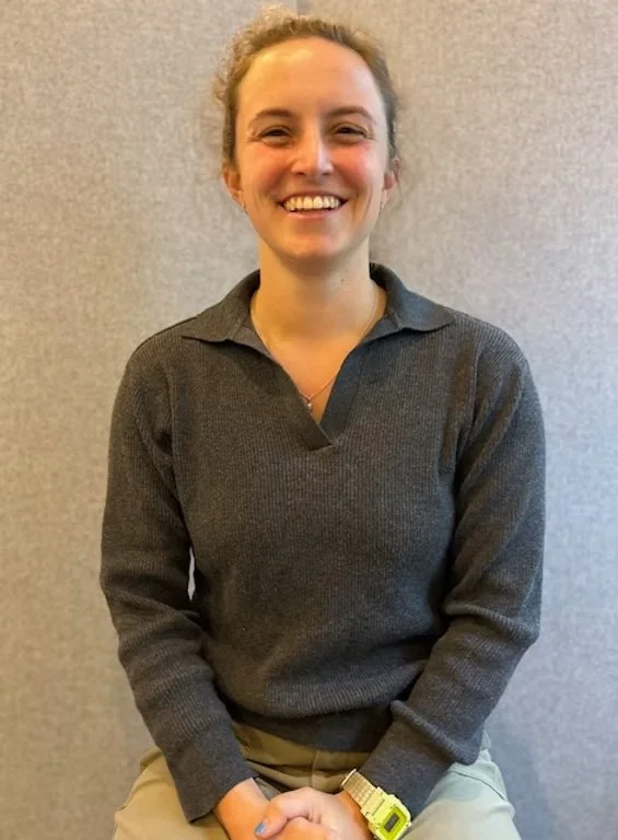 A woman with light skin and curly hair tied back, smiling, wearing a dark gray long-sleeve shirt and beige pants, sitting against a gray wall.