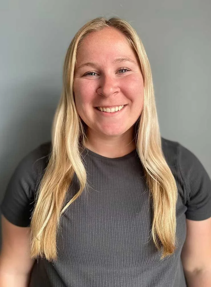 A young woman with long blonde hair smiling, wearing a dark gray t-shirt, standing against a gray background.