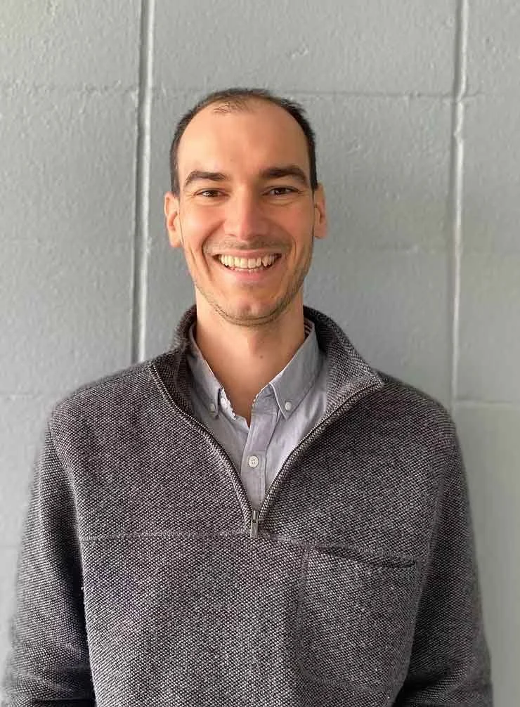 A smiling man with short dark hair and facial stubble, wearing a gray quarter-zip sweater over a light collared shirt, standing against a light gray wall.