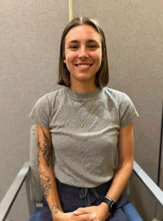 Smiling woman with shoulder-length brown hair, wearing a gray T-shirt, tattooed arms, a necklace, and a black wristwatch, sitting in a chair against a gray wall.