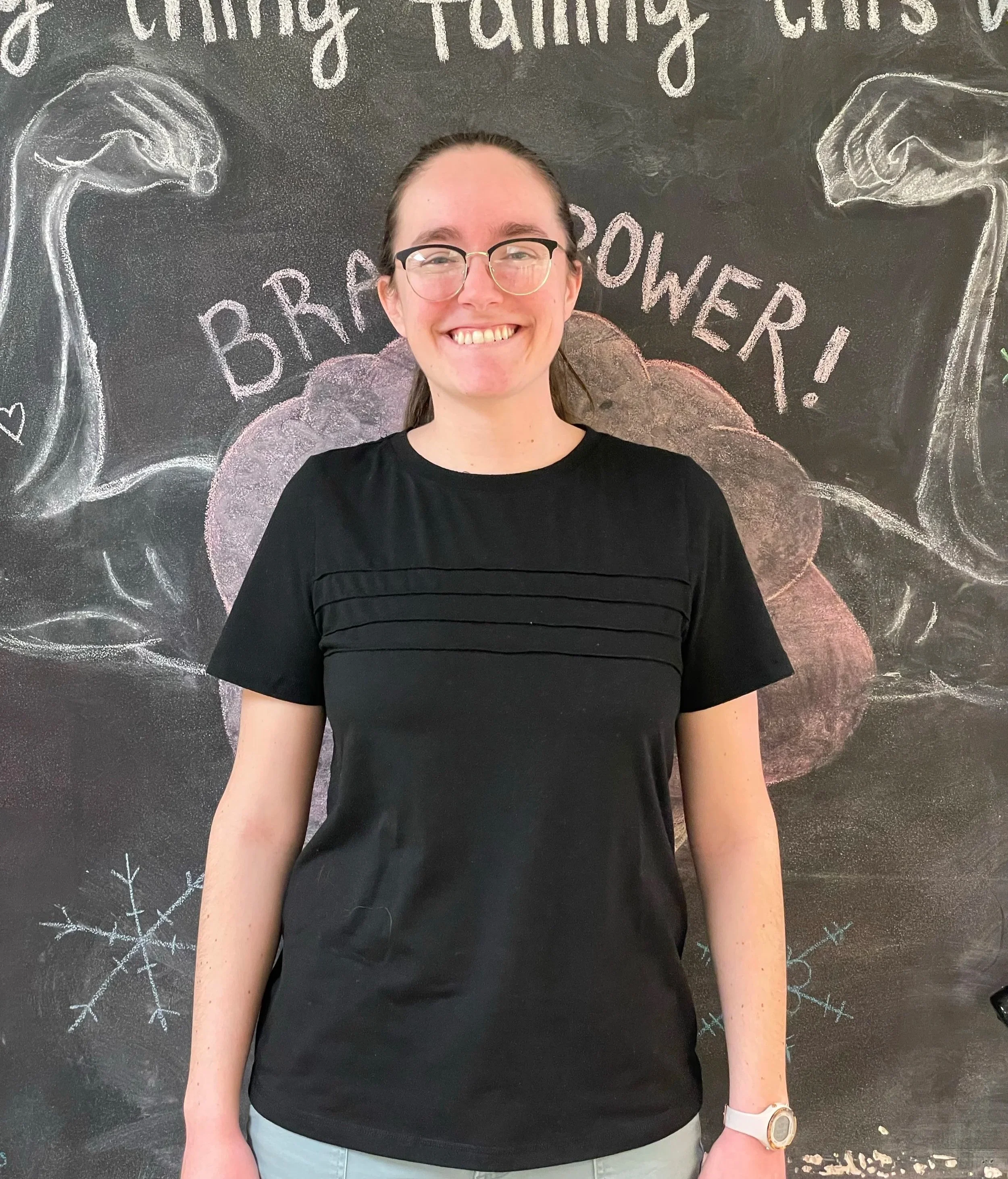 A smiling woman wearing glasses and a black shirt standing in front of a chalkboard wall decorated with festive drawings and the words 'BRRR! POWER!' and 'Warm Fuzzy Feeling' written on it.