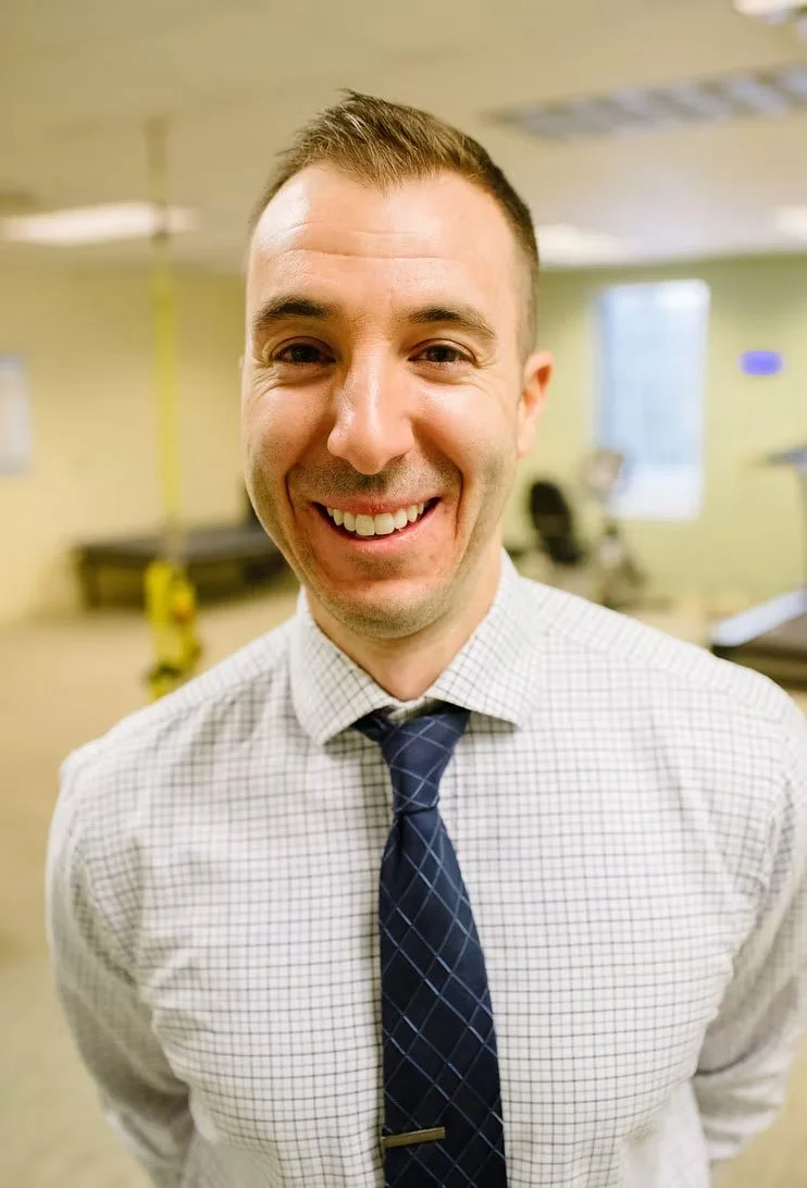 Portrait of a smiling man wearing a checked white shirt and a dark blue tie, standing in an office setting with blurred background.