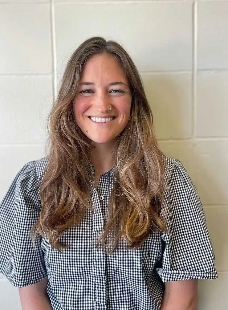 A young woman with long, wavy brown hair smiling in front of a beige brick wall, wearing a black and white checkered blouse and a small nose ring.