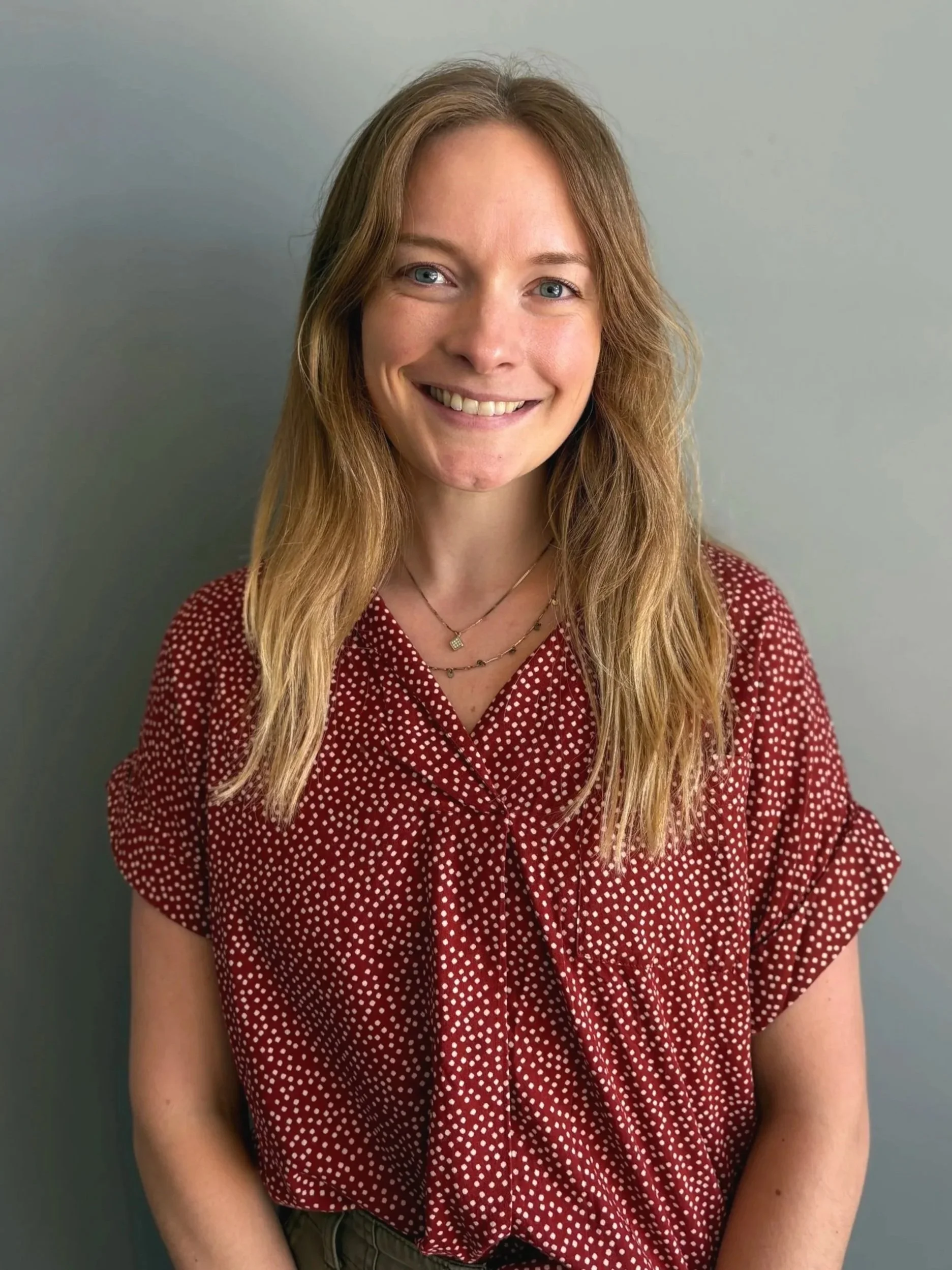 A young woman with light hair, wearing a light gray long-sleeve shirt, smiling and standing against a plain light gray wall.