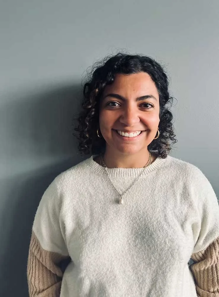 A woman with curly black hair smiling, wearing a cream-colored sweater and gold jewelry, standing against a plain gray background.